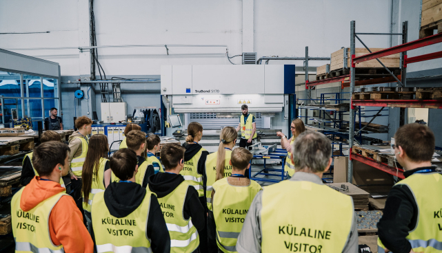Trobikond erkkollaste helkurvestidega noortest tehases. Taga paistab tehase tehnikat ja üks mees, kes tuuri veab.